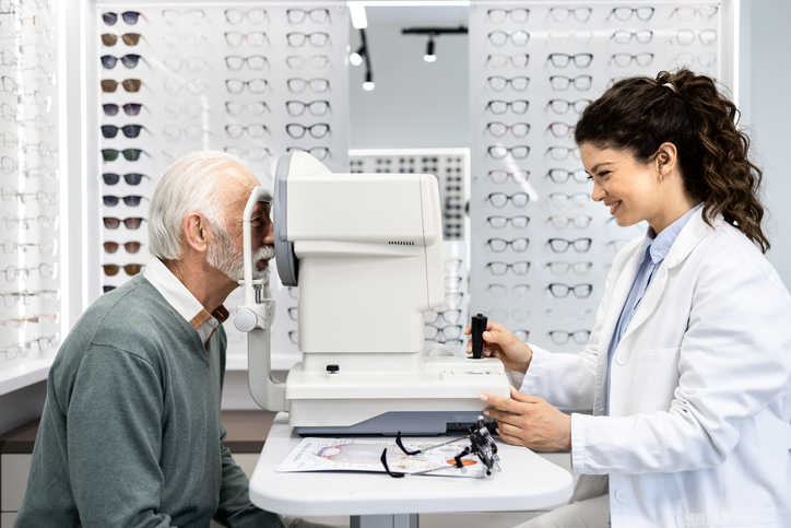 Experienced optometrist checking senior man eyesight focus and diopter using test eyeglasses instrument and eye chart.